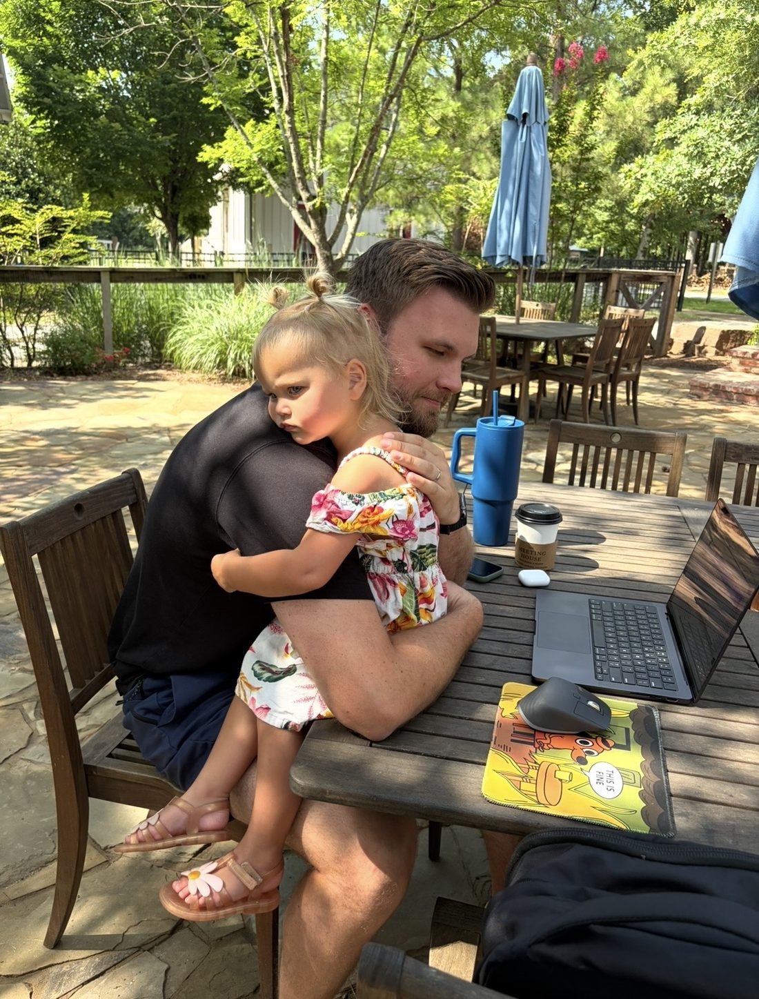 Cameron working on a laptop outside with his daughter asleep against his chest
